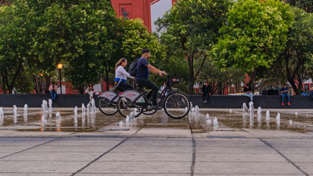 Two cyclists ride past fountains in a vibrant Mexico City park, surrounded by lush greenery and urban architecture.