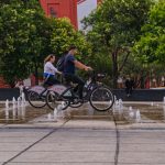 Two cyclists ride past fountains in a vibrant Mexico City park, surrounded by lush greenery and urban architecture.
