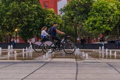 Two cyclists ride past fountains in a vibrant Mexico City park, surrounded by lush greenery and urban architecture.