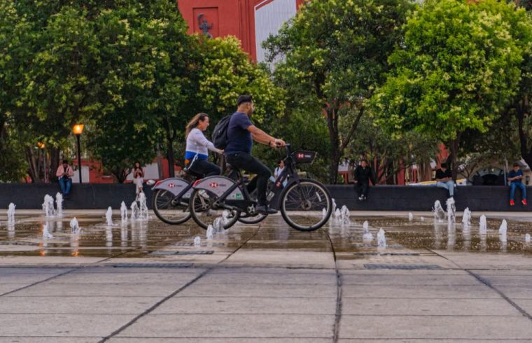 Two cyclists ride past fountains in a vibrant Mexico City park, surrounded by lush greenery and urban architecture.